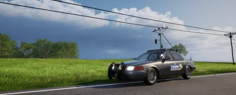 A liveried sedan chase vehicle with roof-mounted instruments, driving a tree-lined rural road